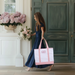 A woman in a blue dress carrying a pink tote bag is walking past a green door with a vase of pink hydrangeas on the windowsill.