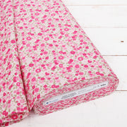 A roll of fabric with a pink floral print pattern is displayed on a white wooden surface.