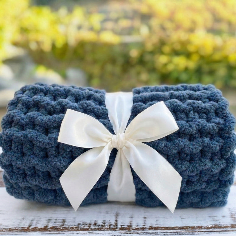 A blue crocheted blanket with a white ribbon bow tied around it, placed on a wooden surface against a blurred background of greenery.