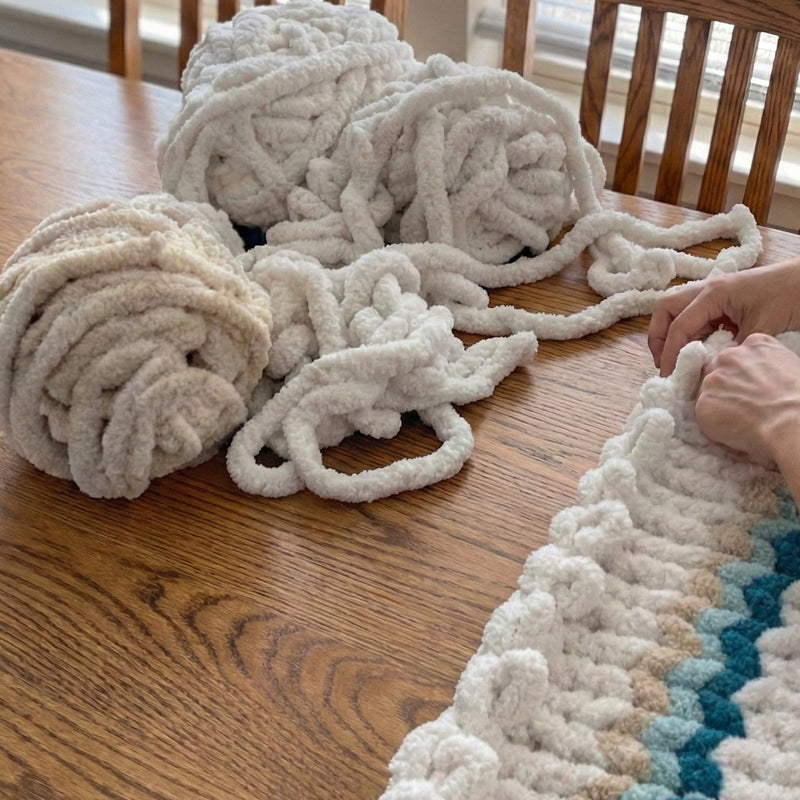 A person's hands are working on a crocheted blanket or rug, with several balls of yarn in various colors and textures scattered on a wooden table.