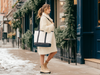 A woman in a white sweater and boots walks down a snowy sidewalk, carrying a large white tote bag.