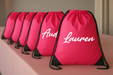 A row of pink drawstring backpacks with the name "Lauren" printed on them, sitting on a pink surface.