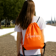 A woman with long brown hair is walking on a brick path, wearing a white shirt and carrying an orange backpack with the name "Audrey" printed on it.