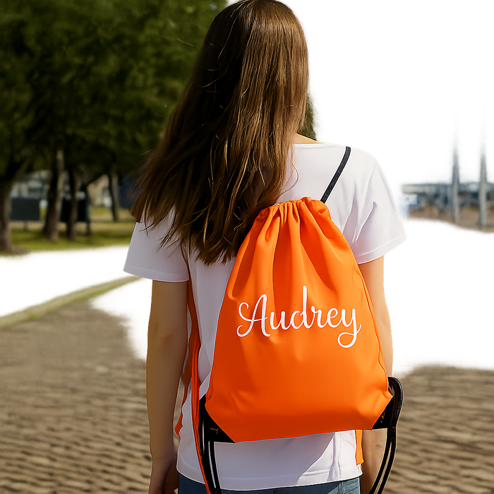 A woman with long brown hair is walking on a brick path, wearing a white shirt and carrying an orange backpack with the name "Audrey" printed on it.