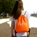A woman with long brown hair is walking on a brick path, wearing a white shirt and carrying an orange backpack with the name "Audrey" printed on it.